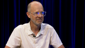 A man with glasses and a white shirt sits in front of blue curtains, looking to the side and smiling slightly.