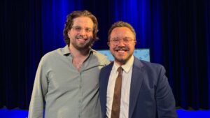 Two men stand side by side, smiling at the camera in front of a blue curtain. One wears a suit and tie, the other a casual shirt.