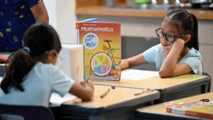 Two young students sit at desks, writing in notebooks with a grade 4 mathematics textbook between them.