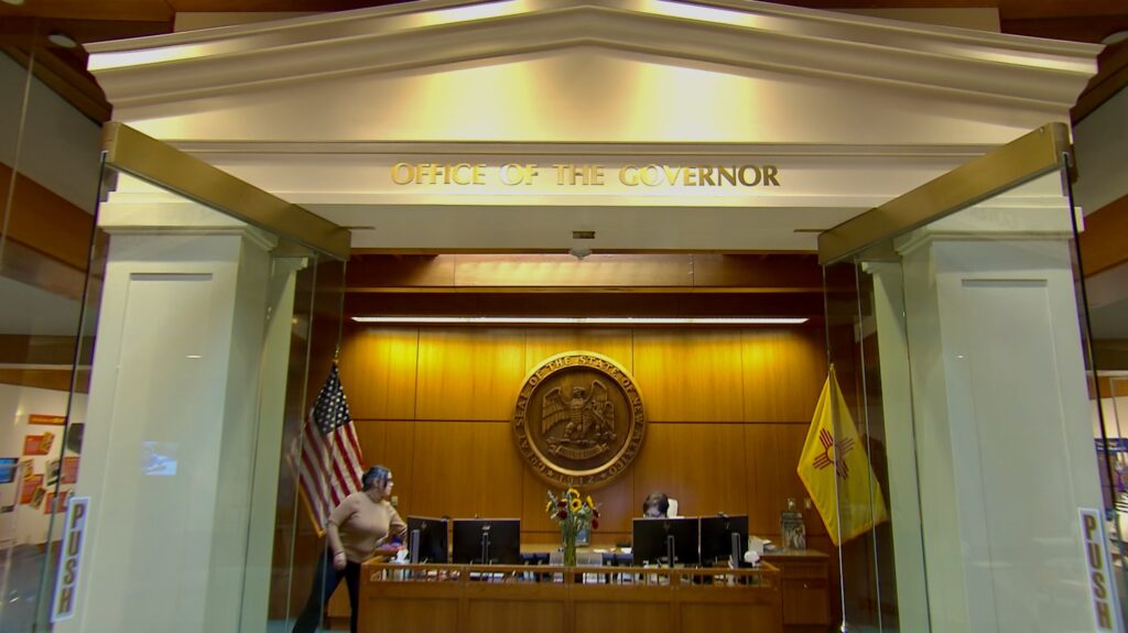 Reception area inside the Office of the Governor with the New Mexico state flag, U.S. flag, and state seal on a wood-paneled wall behind a desk.