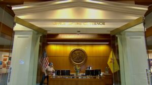 Reception area inside the Office of the Governor with the New Mexico state flag, U.S. flag, and state seal on a wood-paneled wall behind a desk.
