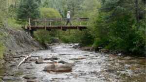 A person walks across a wooden footbridge spanning a rocky mountain stream.