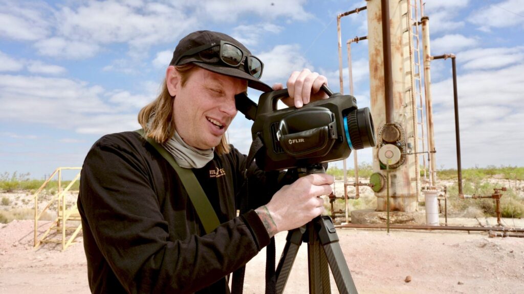 A person in a baseball cap using a FLIR thermal imaging camera on a tripod at an industrial site.