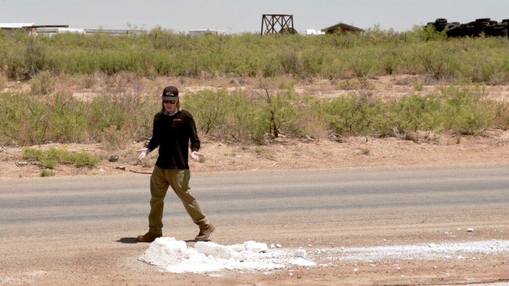 A person wearing a cap and long-sleeved shirt walks along the shoulder of a road near a pile of white, chalky material.
