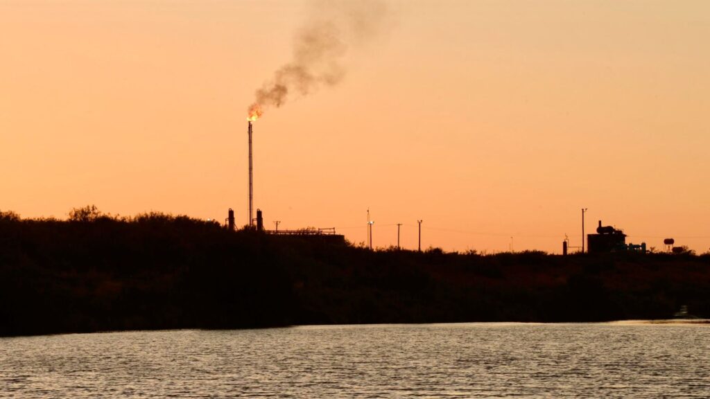 An industrial flare stack emitting smoke against a sunset sky above a body of water.