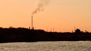 An industrial flare stack emitting smoke against a sunset sky above a body of water.