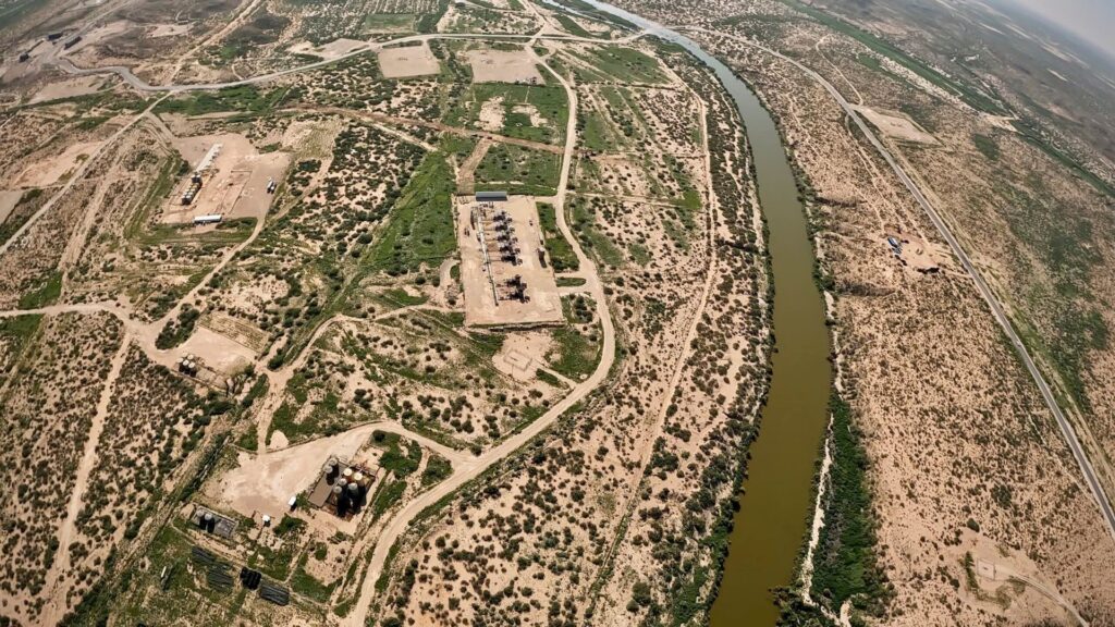 Aerial view of oil field infrastructure, including pump jacks and storage tanks, situated on arid land beside a winding river.