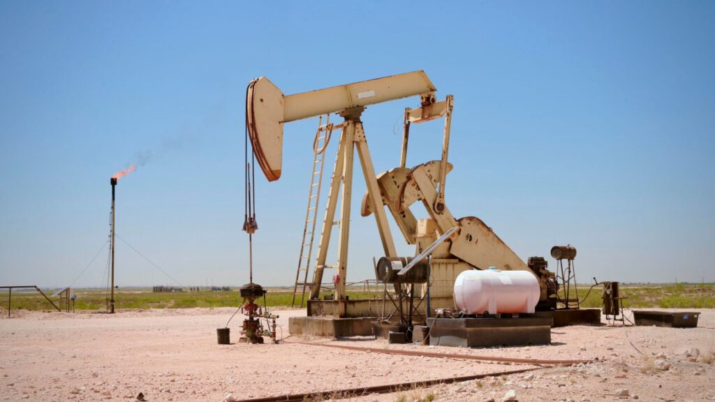 An oil pump jack operates in a desert landscape with a gas flare burning in the distance.