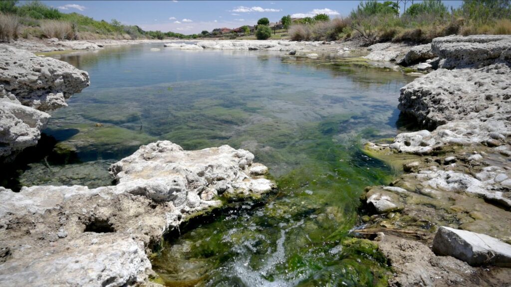 A stream with clear water flows over mossy rocks into a shallow pool surrounded by limestone banks.