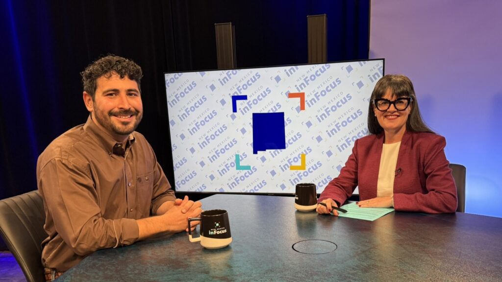 A man and a woman sit at a desk in a television studio in front of a monitor displaying the “New Mexico inFocus” logo.