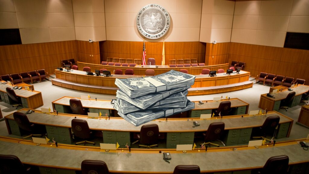 A large stack of cartoon-style paper money resting in the center of an empty New Mexico legislative chamber.