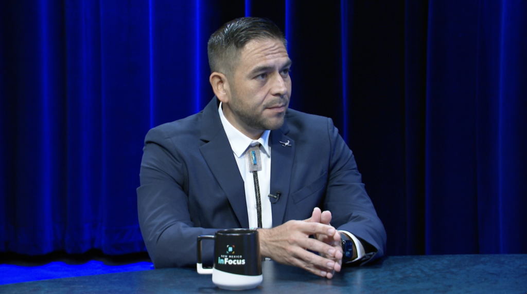 A man in a suit and bolo tie sits at a table with a “New Mexico InFocus” mug in front of a blue curtain.