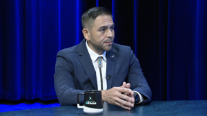 A man in a suit and bolo tie sits at a table with a “New Mexico InFocus” mug in front of a blue curtain.