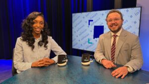 Two news anchors sit at a desk with “New Mexico InFocus” mugs in front of a blue background screen.