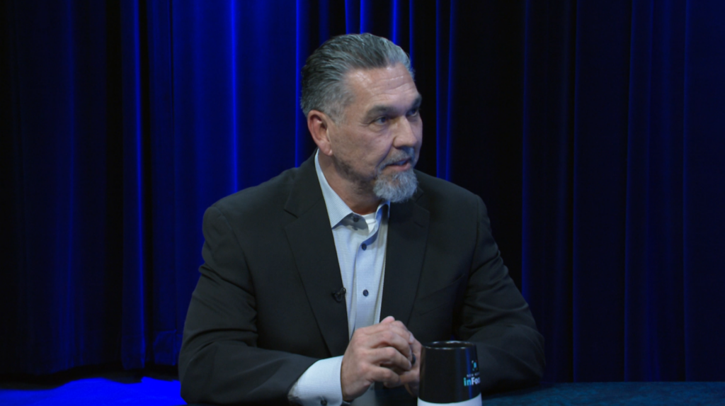 A man with a goatee wearing a suit jacket and dress shirt sits at a table in front of blue curtains.