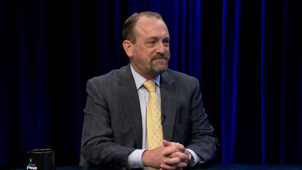 A man in a suit and yellow tie sits at a table with his hands clasped, in front of a dark blue curtain background.