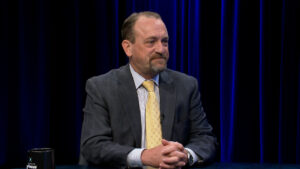 A man in a suit and yellow tie sits at a table with his hands clasped, in front of a dark blue curtain background.