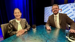 Two people sit at a blue table with coffee mugs, facing the camera, on a studio set with screens displaying the words “in Focus” in the background.
