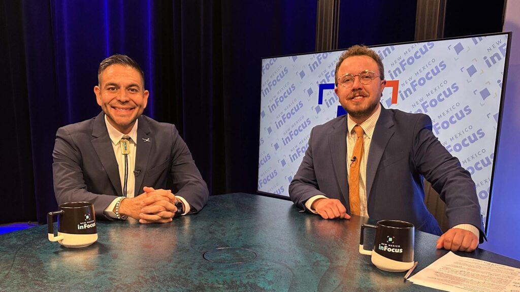Two men in suits sit at a news desk in front of a New Mexico InFocus branded screen, each with a matching branded mug in front of them.