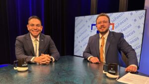 Two men in suits sit at a news desk in front of a New Mexico InFocus branded screen, each with a matching branded mug in front of them.
