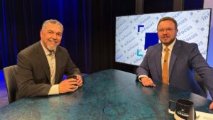 Two men in suits sitting at a desk in a television studio in front of a monitor displaying the New Mexico InFocus logo.