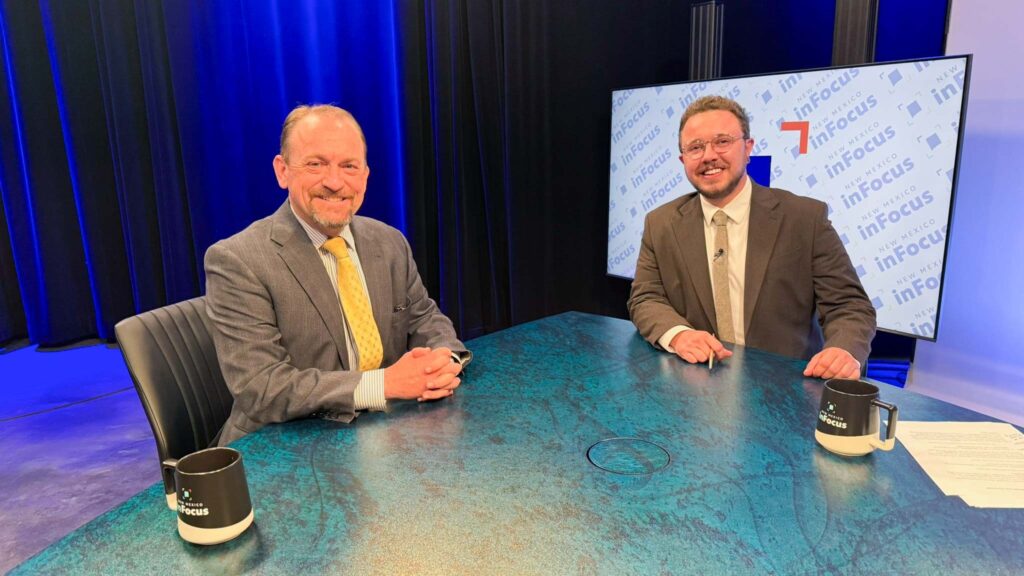 Two men in suits sit at a teal table in a TV studio with “in focus” on a screen behind them. Two branded mugs and papers are on the table. The background features dark curtains and blue lighting.