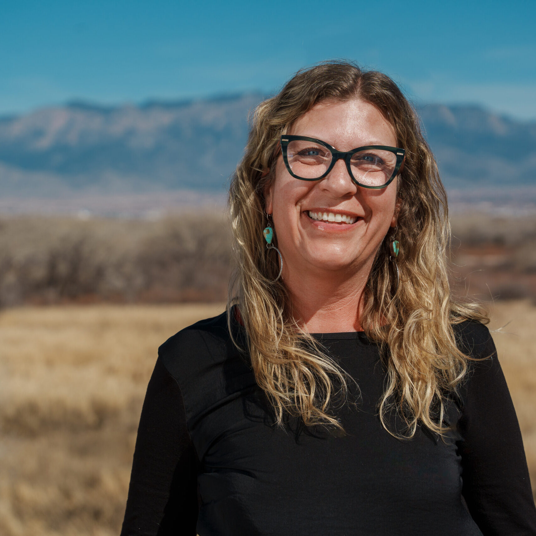 Smiling woman with glasses standing outdoors with a mountainous backdrop.