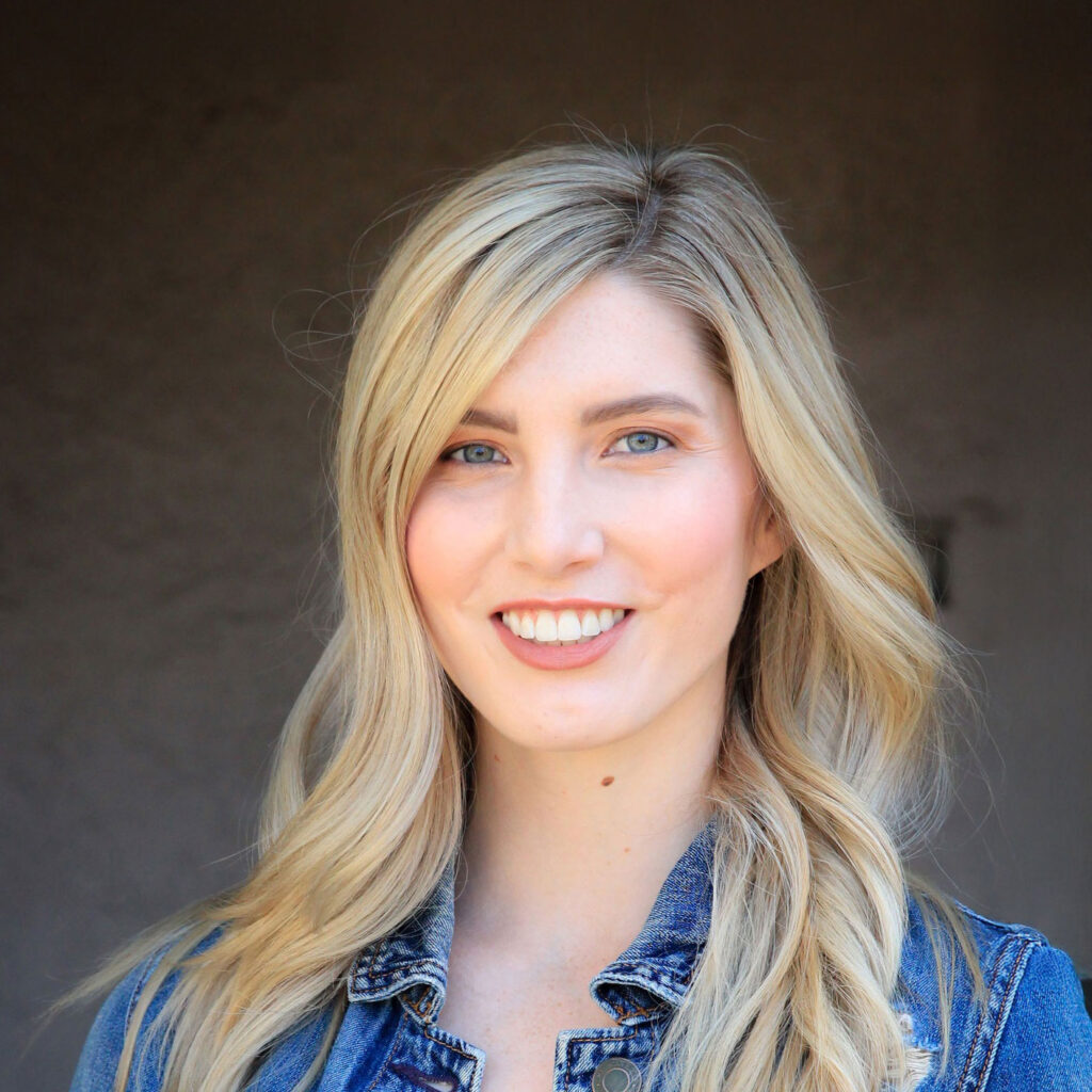 CailleyApril2018-6912 A woman with long blonde hair and blue eyes smiles at the camera, wearing a blue denim jacket against a plain background.