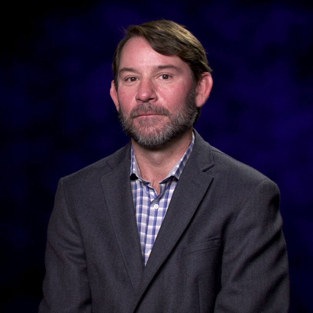 A man with a beard is standing in front of a purple background.