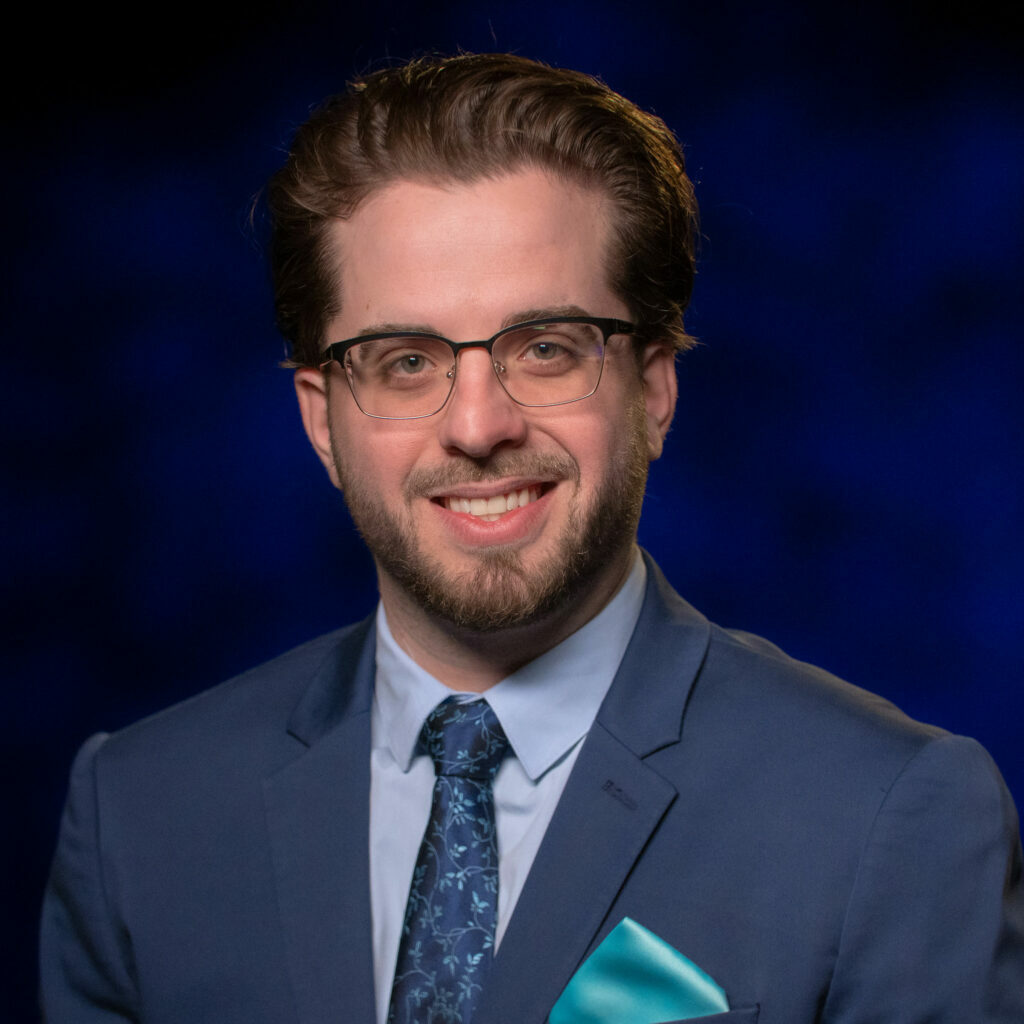 NMPBS-Portraits_Lou-DiVizio(1) A man wearing glasses and a blue tie.