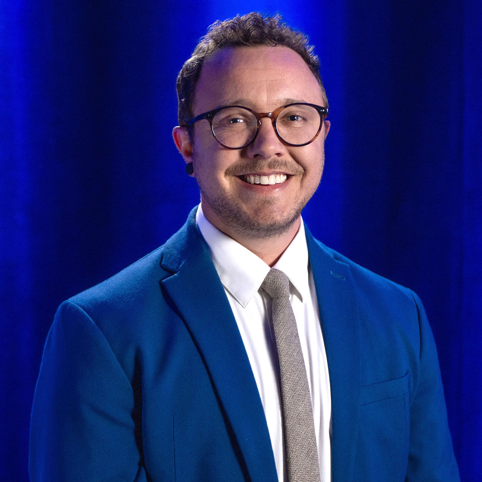 PBS-Headshot-Nash-Jones-Final A person with short brown hair, glasses, and a beard, wearing a blue suit, white shirt, and gray tie, stands smiling in front of a blue curtain background.