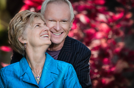 Two older people hold each other and smile in front of a red bush.