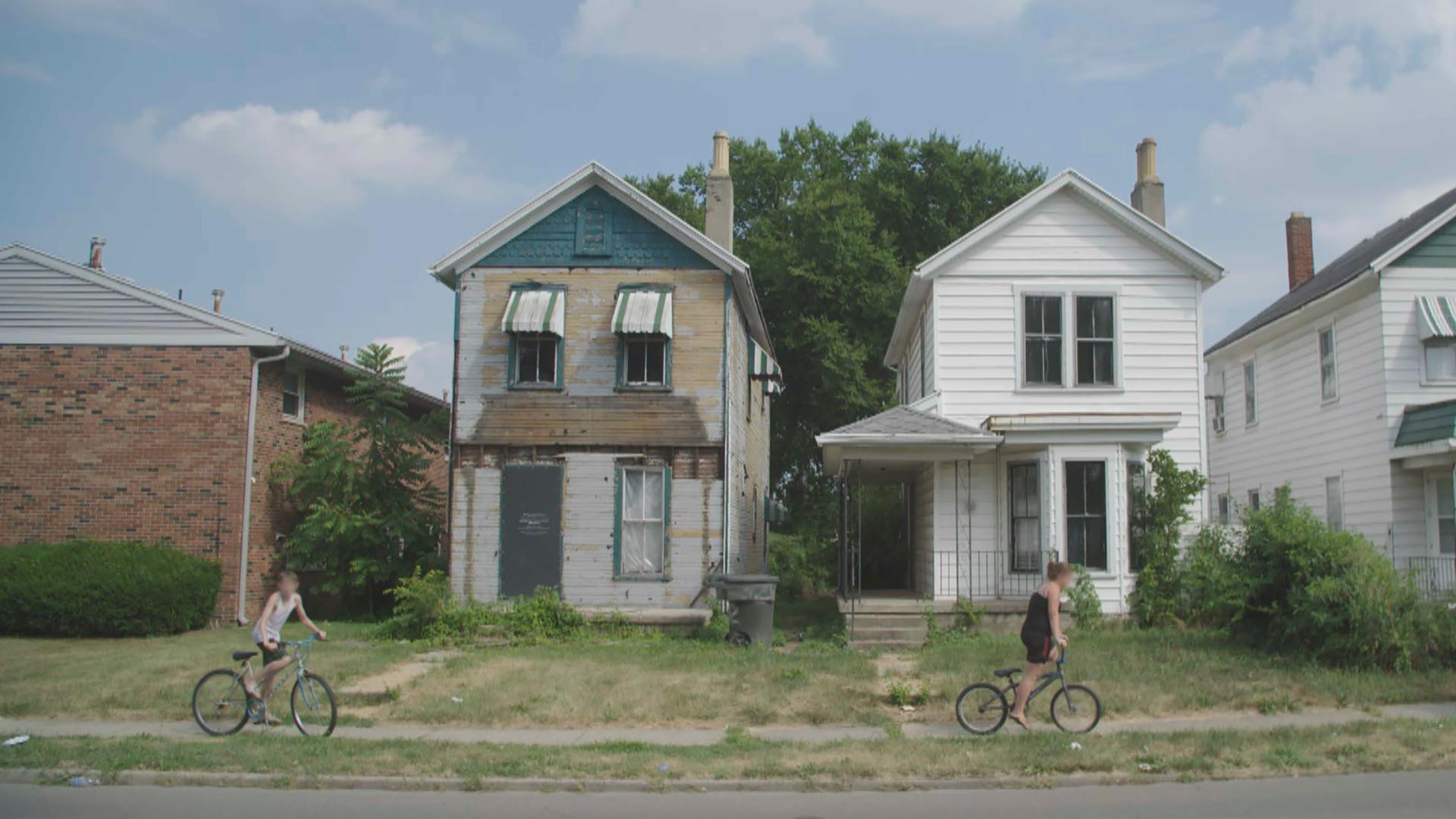 Two people on bicycles ride down a street in front of abandoned houses.