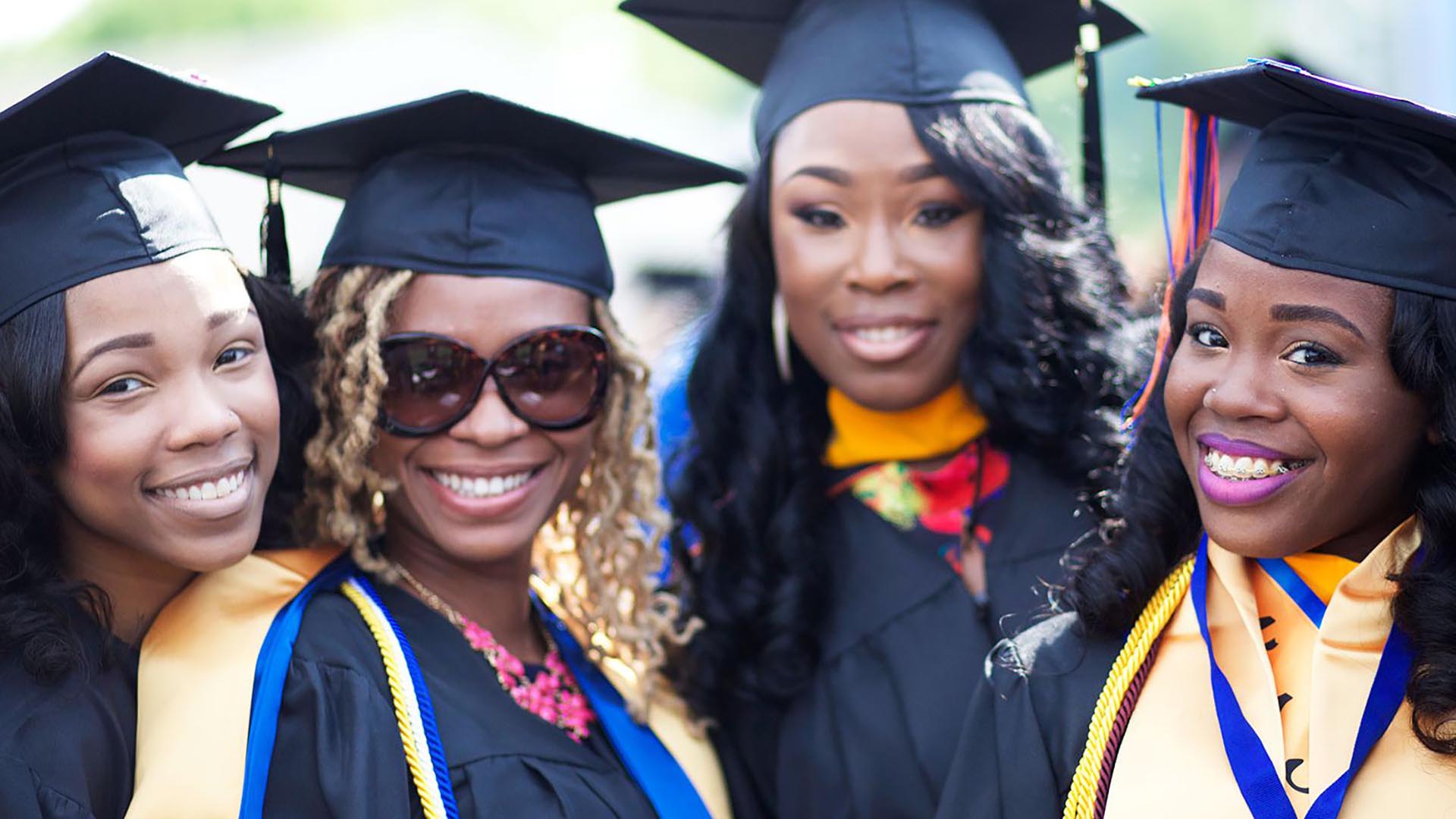 tell-them-we-are-rising Four women smile and pose for the camera in graduation caps and gowns.