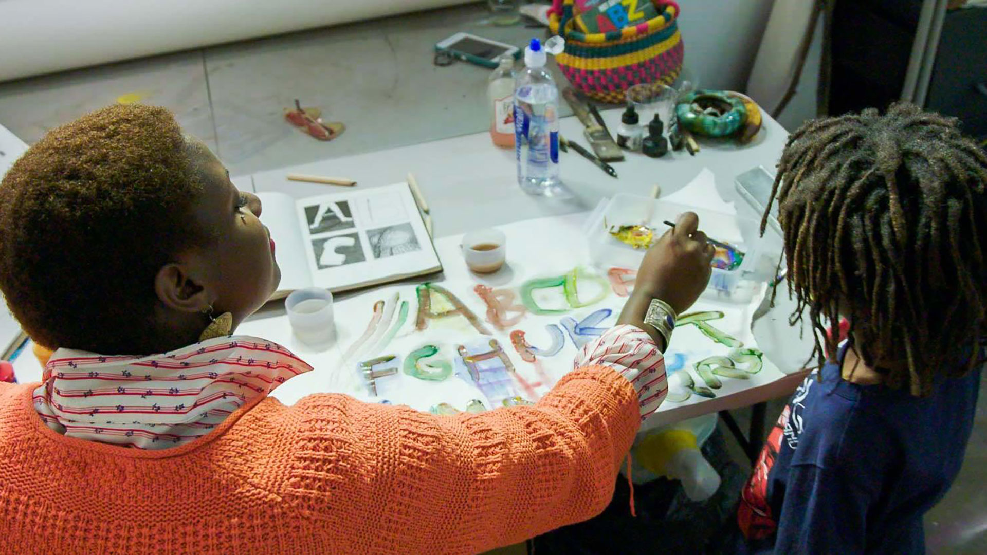 A person dips a brush into paint as a child watches next to them.