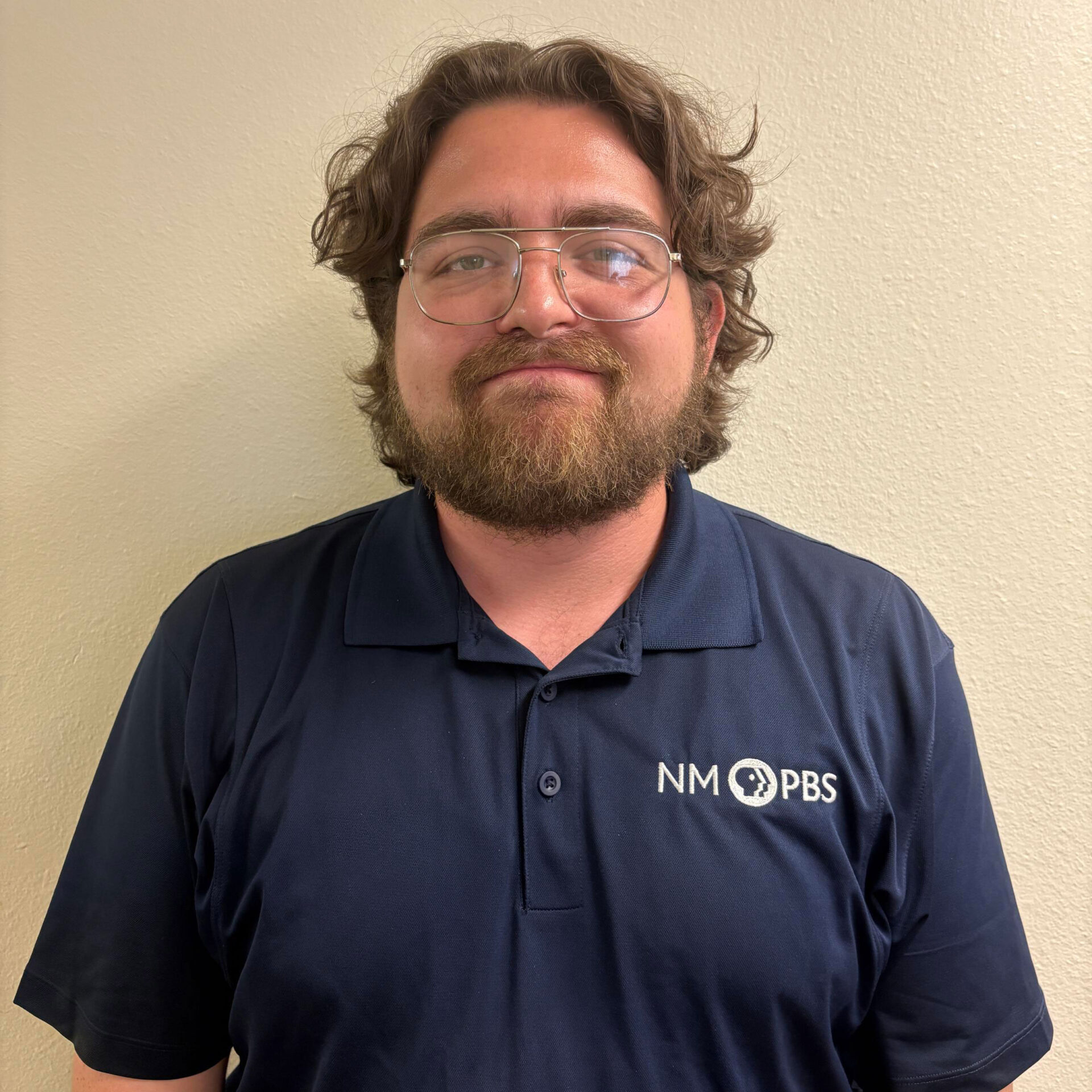 Aiden-Breau A person with glasses and a beard wearing a navy NM PBS polo shirt stands in front of a plain light-colored wall.