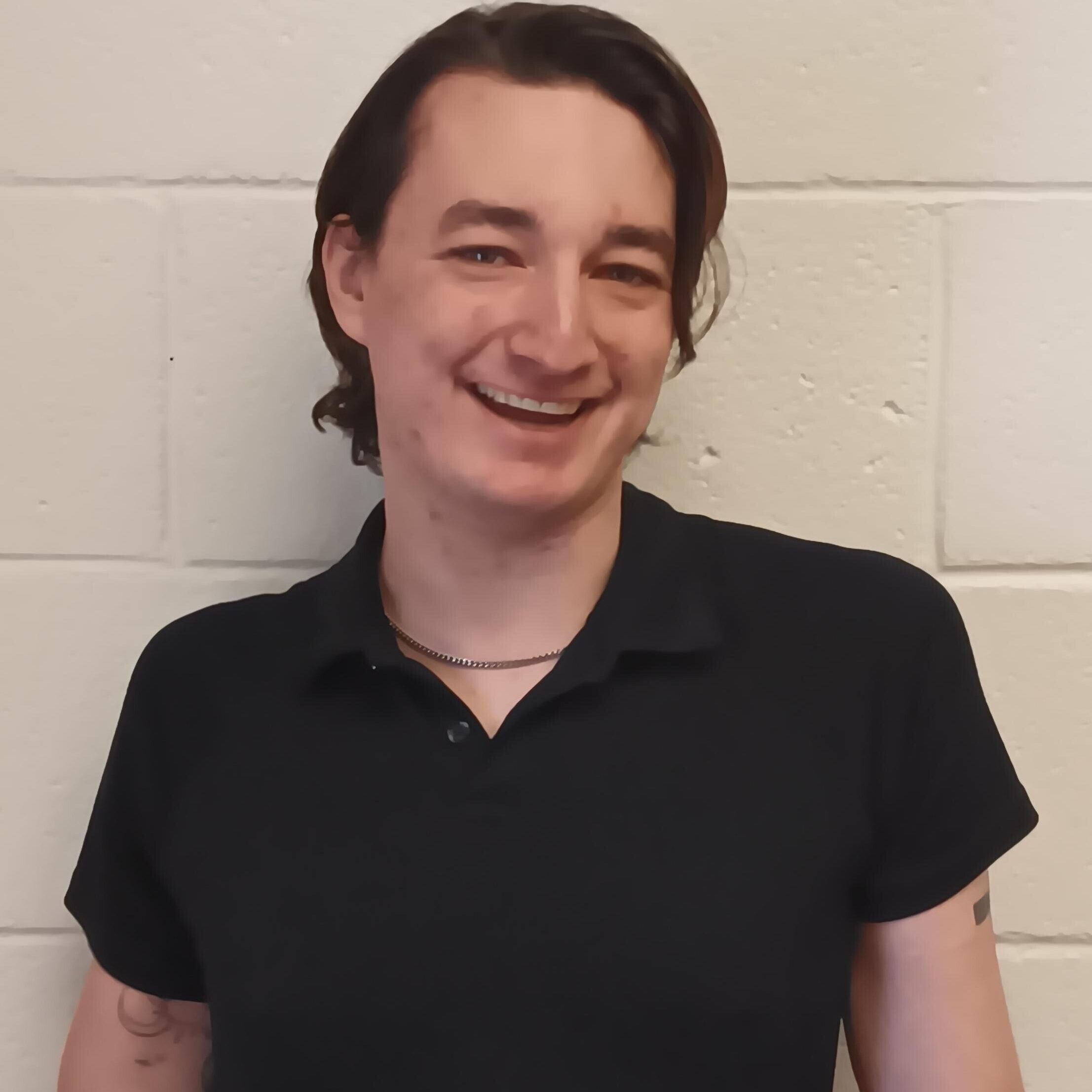 Person with medium-length brown hair, wearing a black polo shirt and a chain necklace, is standing against a light-colored brick wall and smiling at the camera.