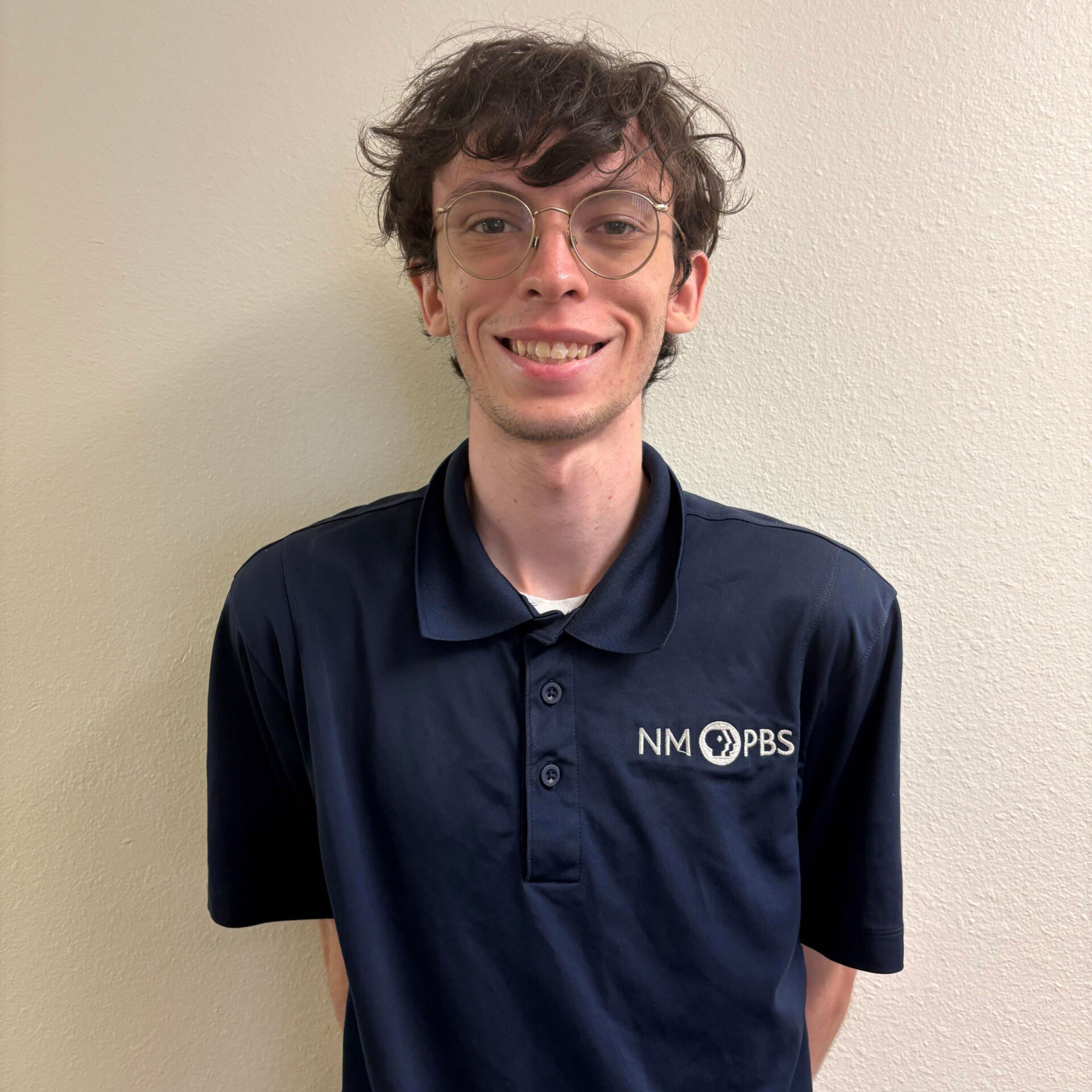 Jacob-Sonney A person wearing glasses and a navy NM PBS polo shirt stands in front of a plain off-white wall, smiling at the camera.