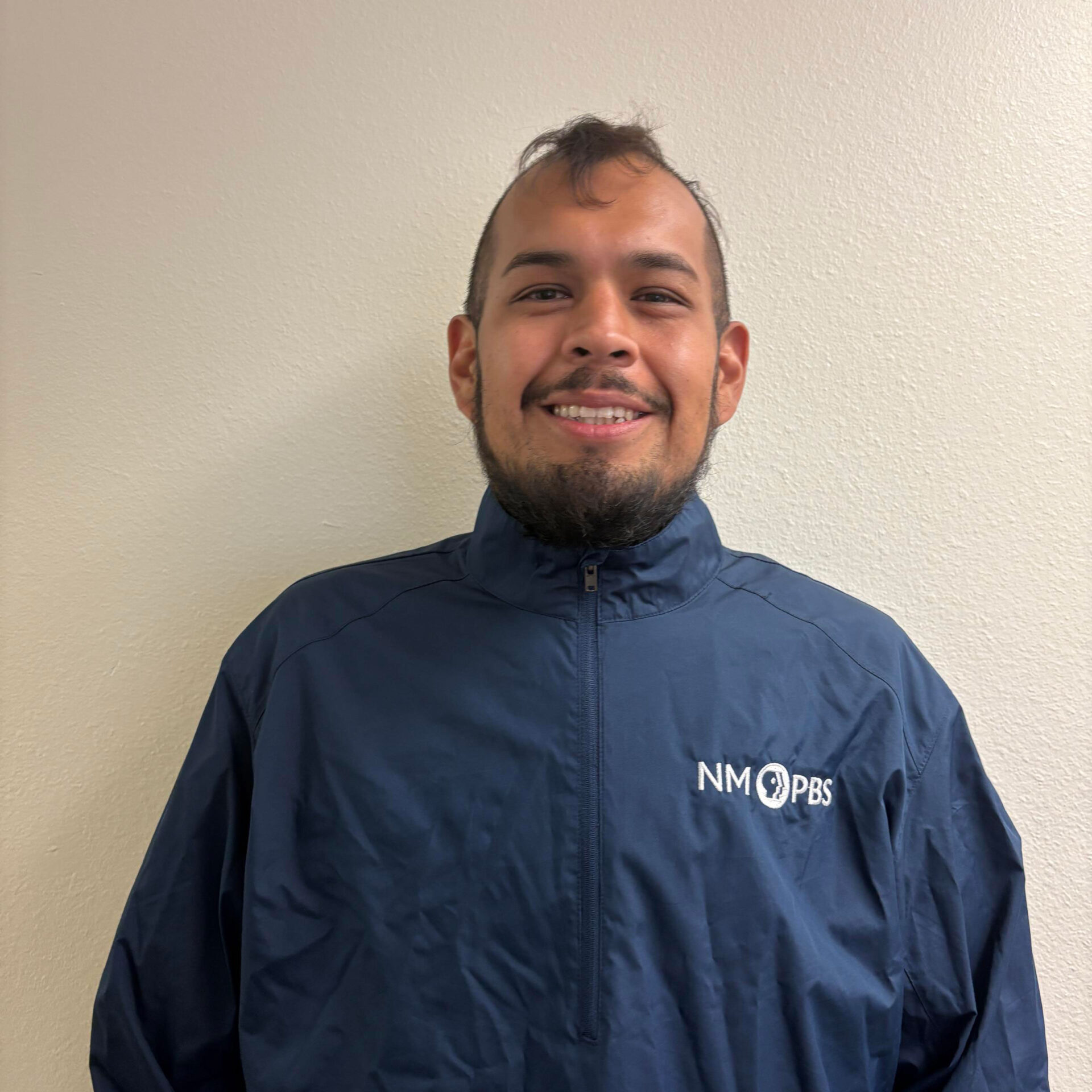Jalen-Sloan A person wearing a navy blue NM PBS jacket stands against a plain, light-colored wall, smiling at the camera.