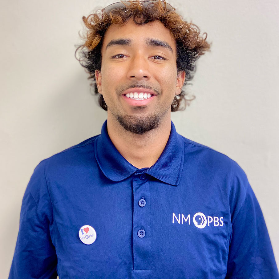 Man wearing a blue NM PBS polo shirt and an “I love Chris” button stands in front of a plain light-colored wall, smiling at the camera.