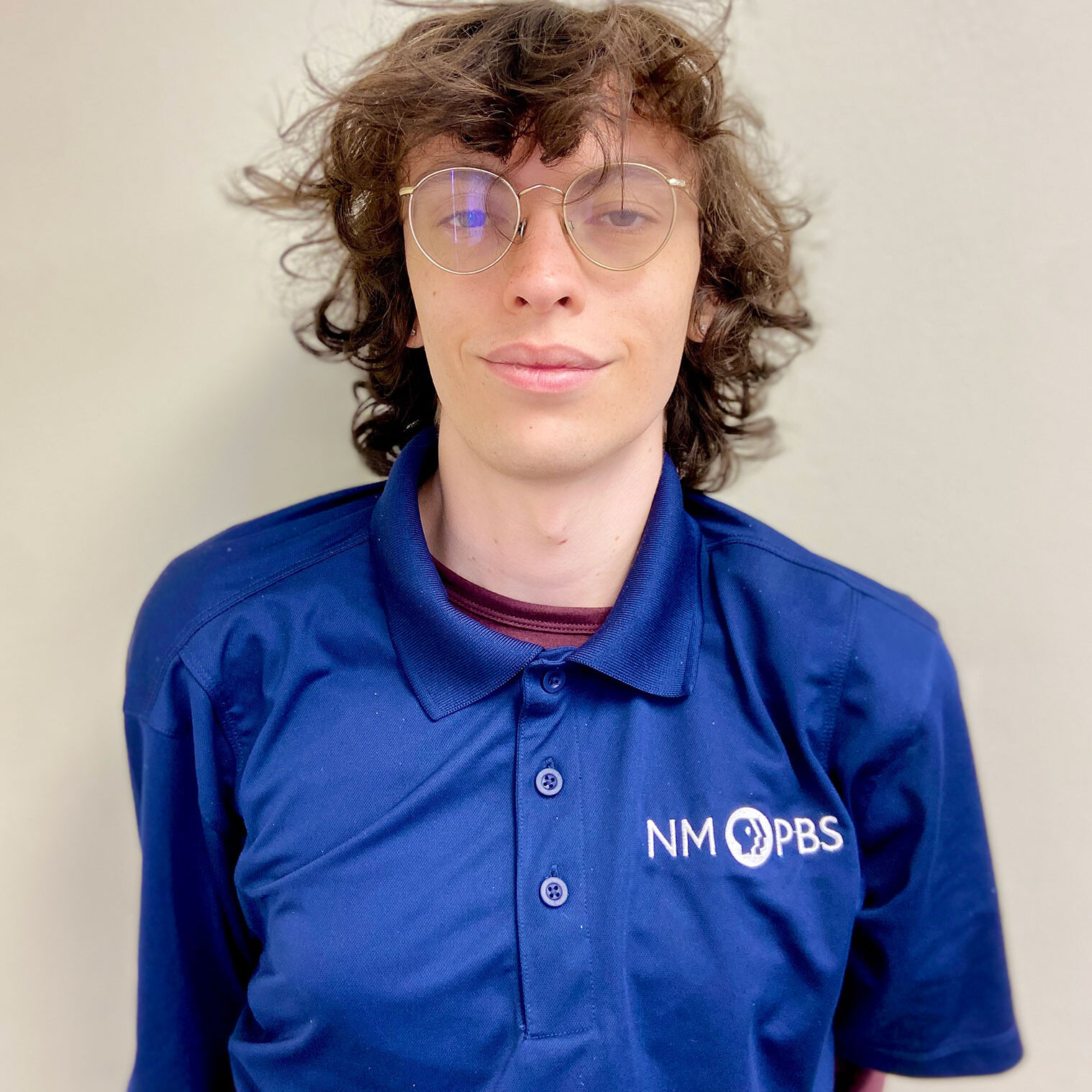 Person with curly hair and glasses wearing a blue NM PBS polo shirt, standing against a plain light-colored wall.