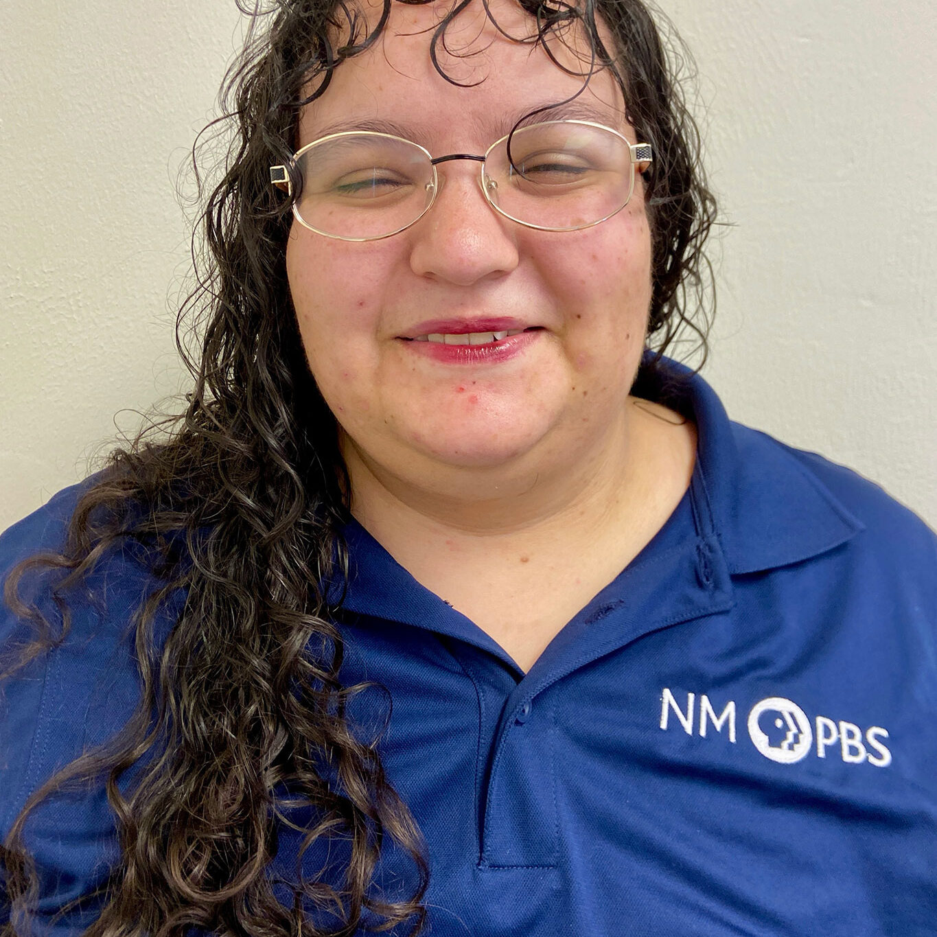 Person with long curly hair and glasses wearing a blue NM PBS polo shirt, smiling in front of a plain light-colored wall.