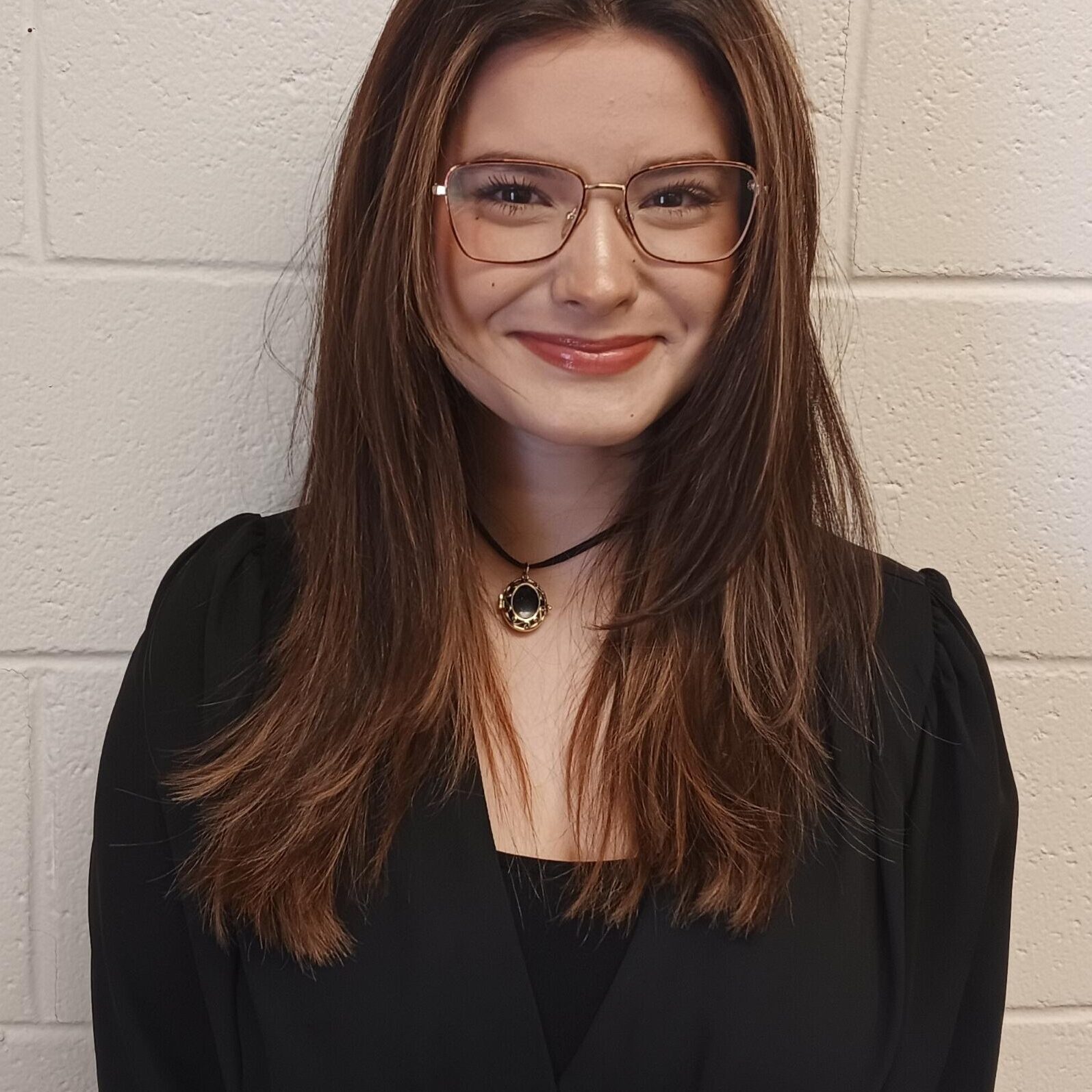 A woman with long brown hair, glasses, and a black outfit stands smiling in front of a white brick wall.