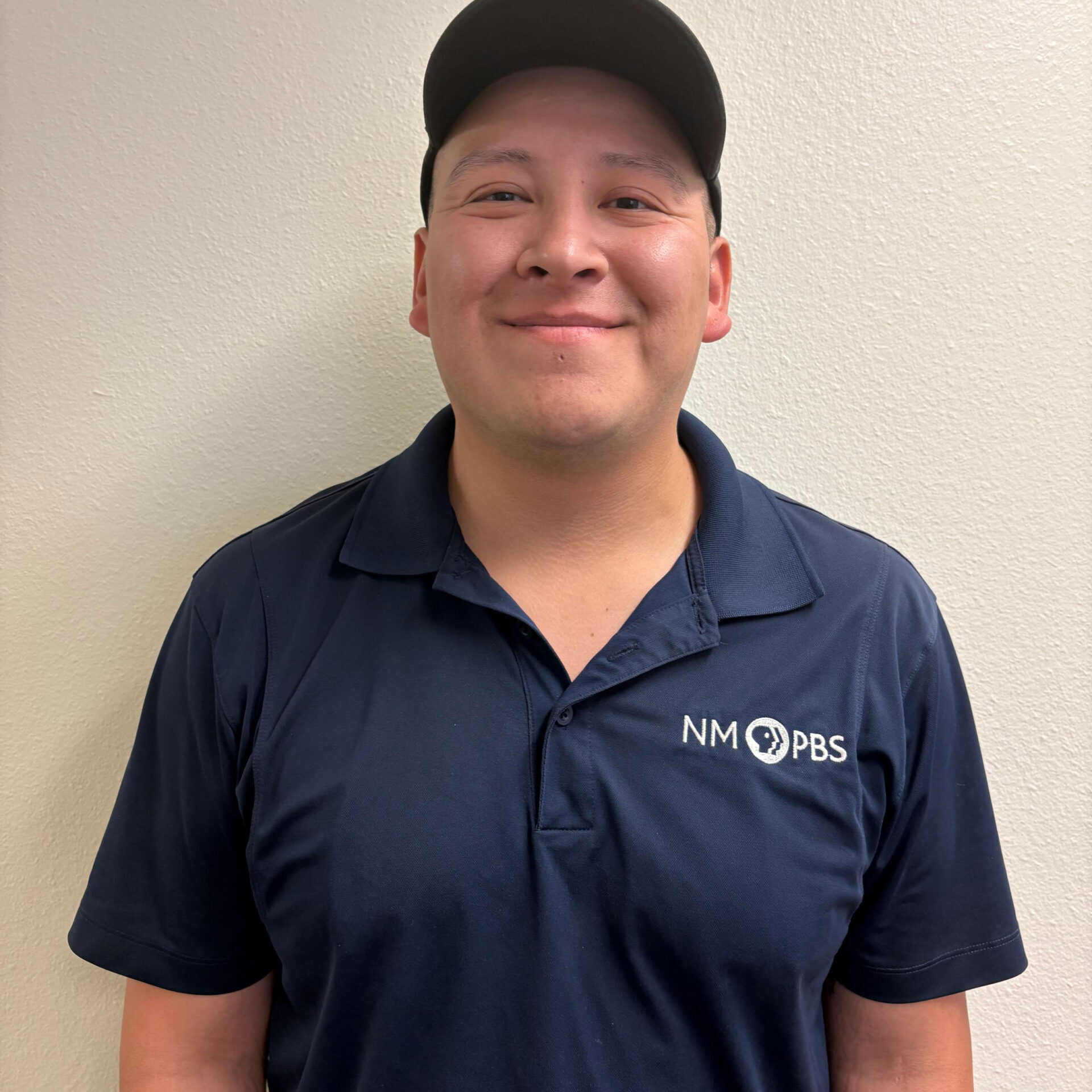 Nicholas-Encinias A person wearing a navy NM PBS polo shirt and a black cap stands against a plain light-colored wall, smiling at the camera.