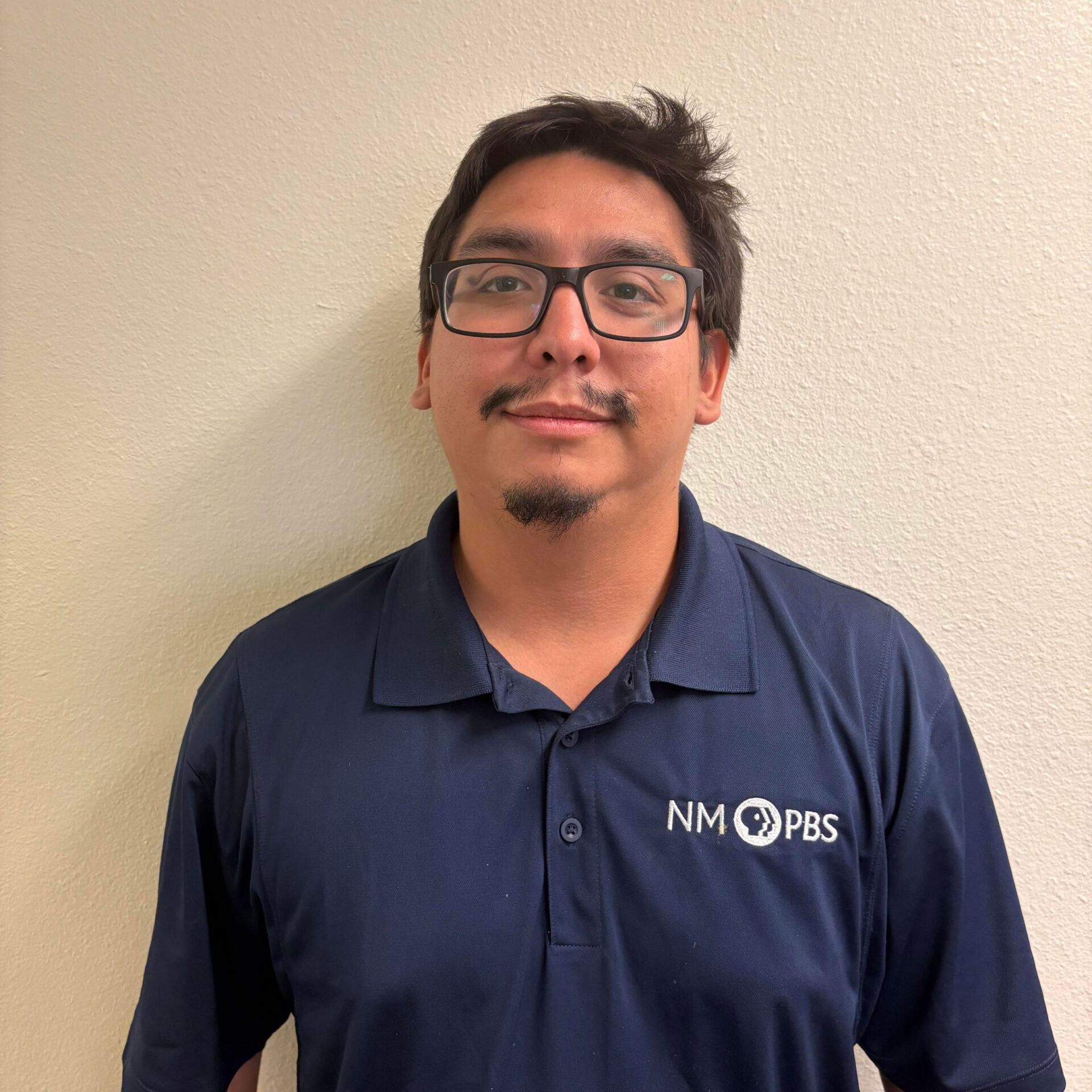 Nick-Dominguez A person with glasses and a mustache wearing a navy blue NM PBS polo shirt stands against a plain light-colored wall.
