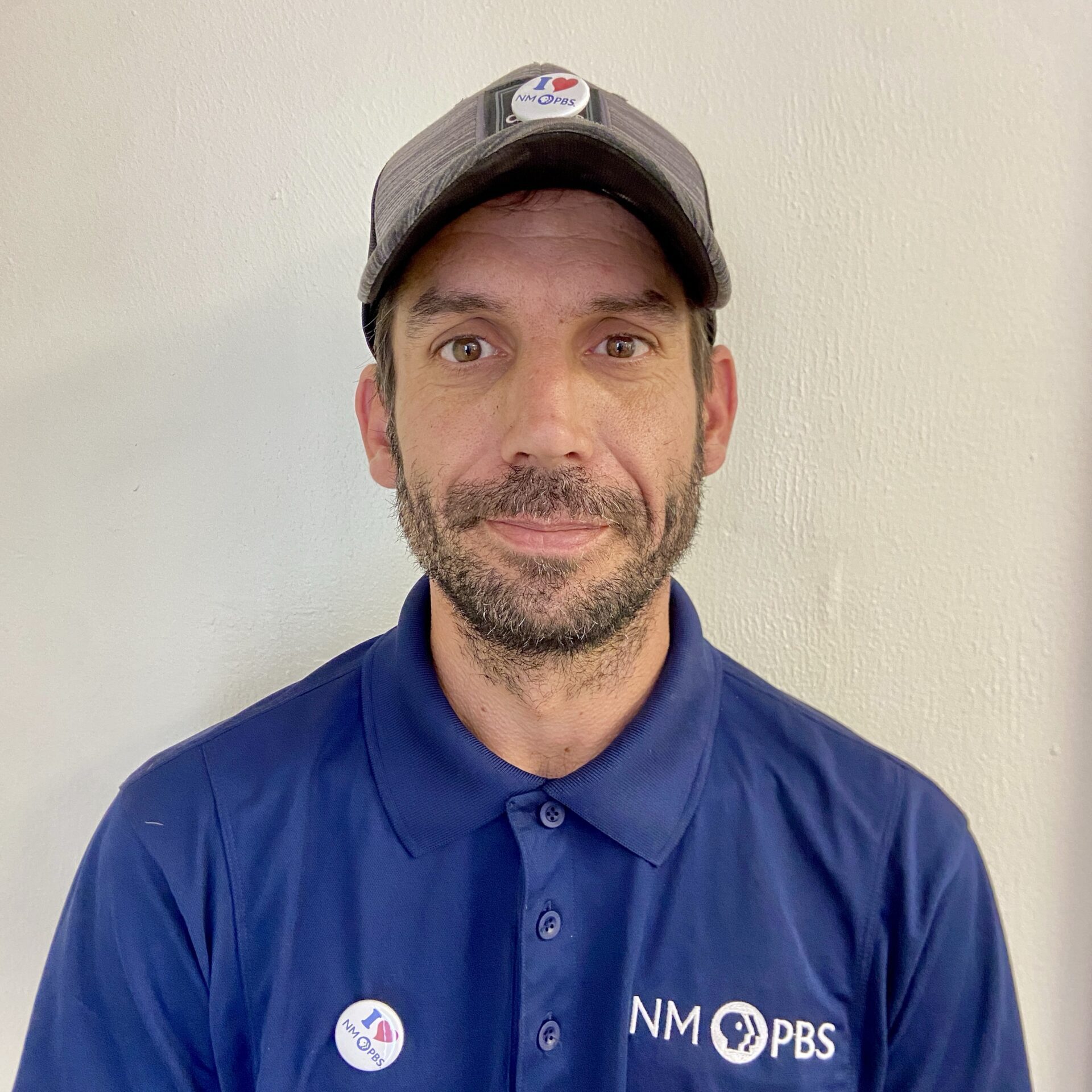 A man wearing a blue NM PBS shirt and cap stands against a light-colored wall. He has a short beard and is wearing two “I Voted” stickers.