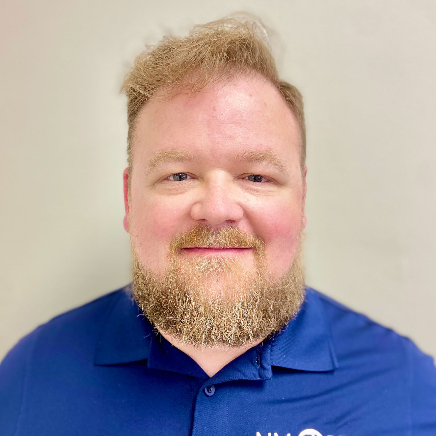 A person with a beard and short hair is wearing a blue NM PBS shirt and standing in front of a plain light-colored background.
