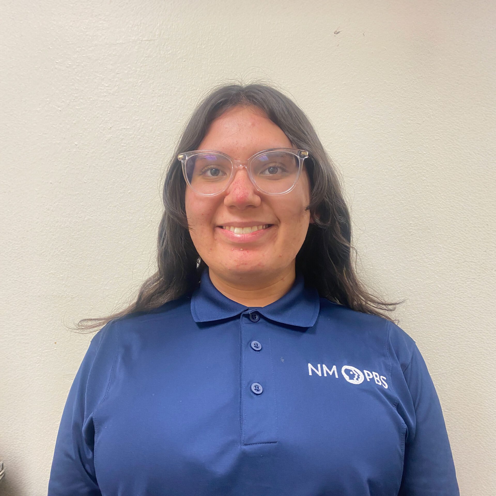 image-(1) A person with long dark hair and glasses wearing a blue NM PBS polo shirt stands in front of a light-colored wall, smiling at the camera.