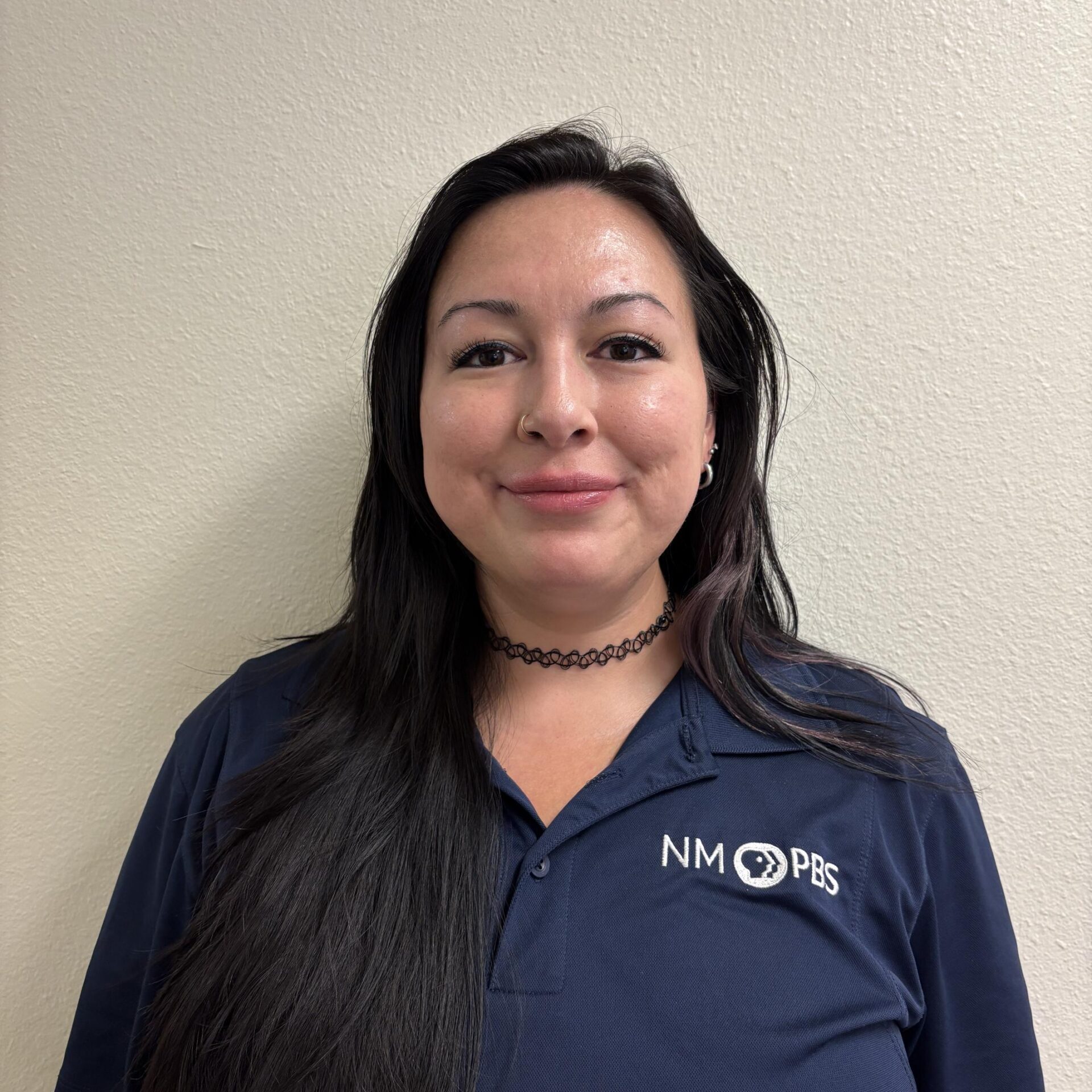 A woman with long dark hair wearing a navy blue NM PBS polo shirt stands against a light-colored wall, smiling softly at the camera.